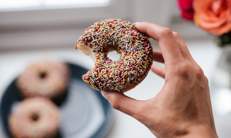 Hand holds a donut with brown icing and colorful sprinkles over a plate of donuts containing a sugar substitute